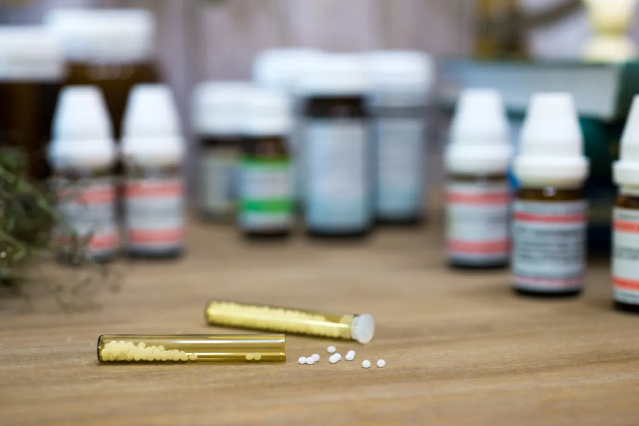 Small vials of white pellets on a wooden table in front of blurred medicine bottles