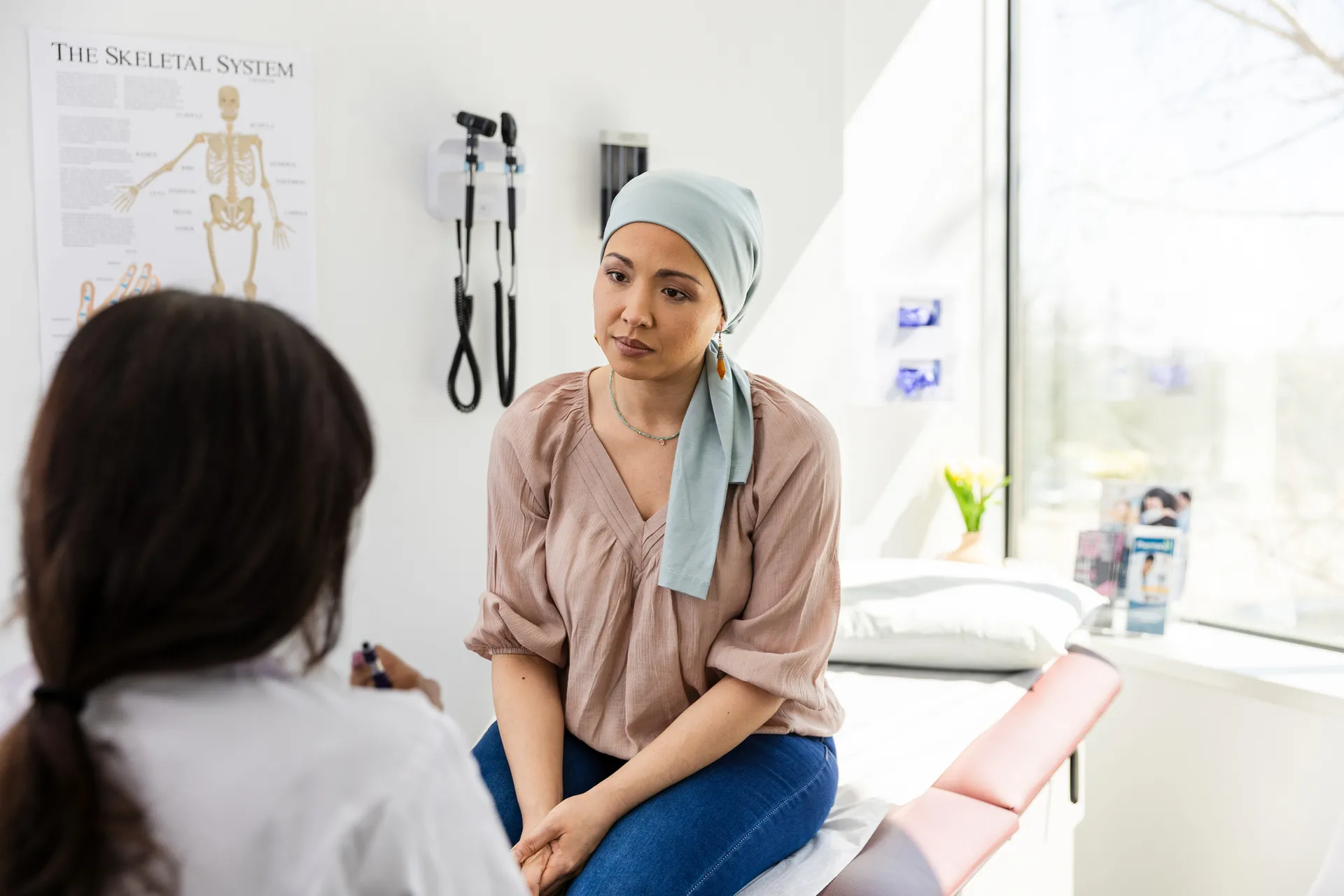 Woman in a headscarf sitting on an exam table talking with a doctor, skeletal system poster on the wall