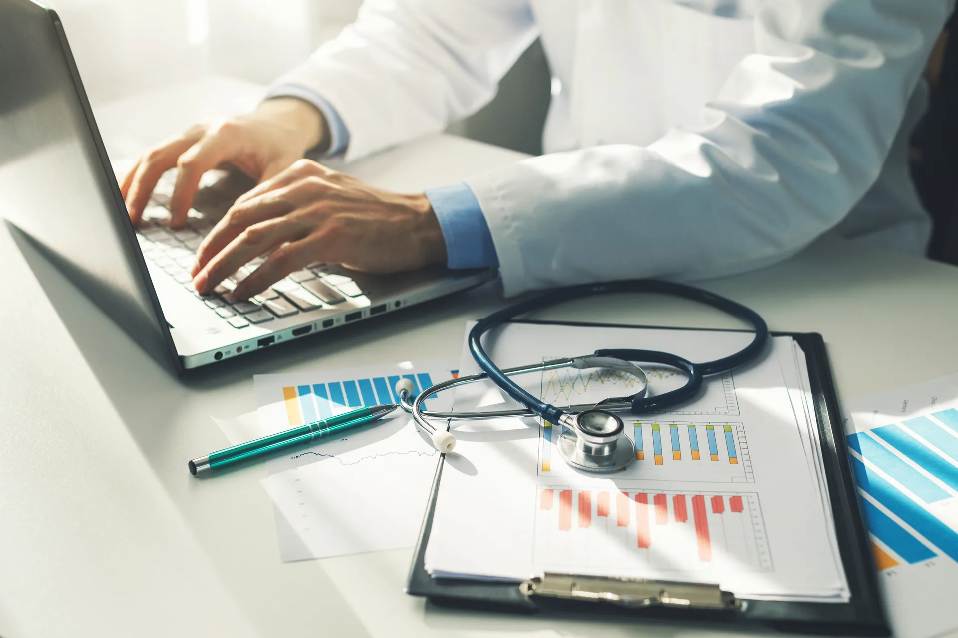 Doctor types on a laptop beside a stethoscope and clipboard of printed charts
