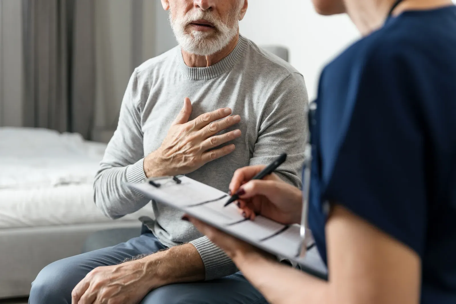 Older man with his hand on his chest describes symptoms to a clinician taking notes on a clipboard