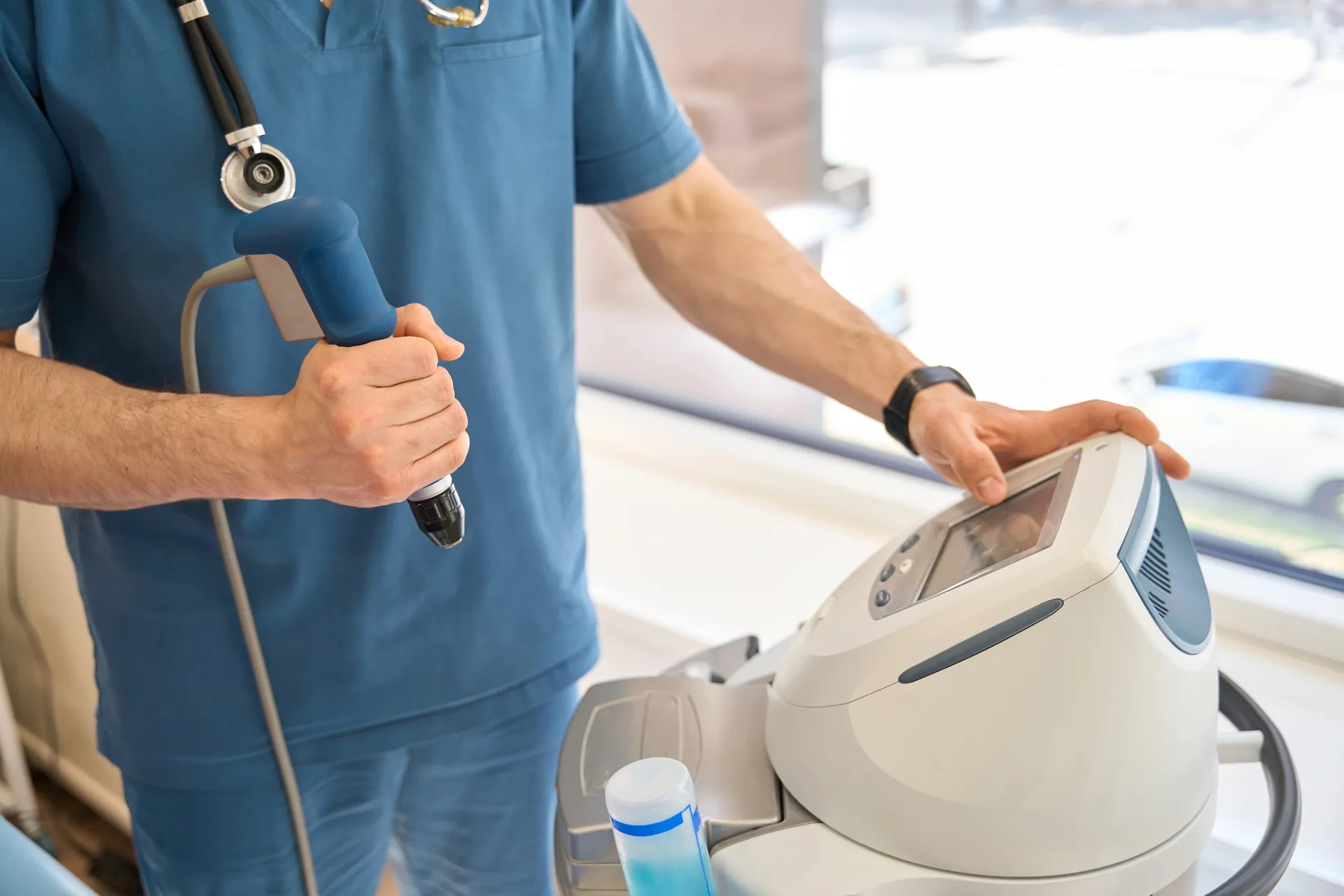 Clinician in scrubs holding the handpiece of a shockwave therapy device next to its console