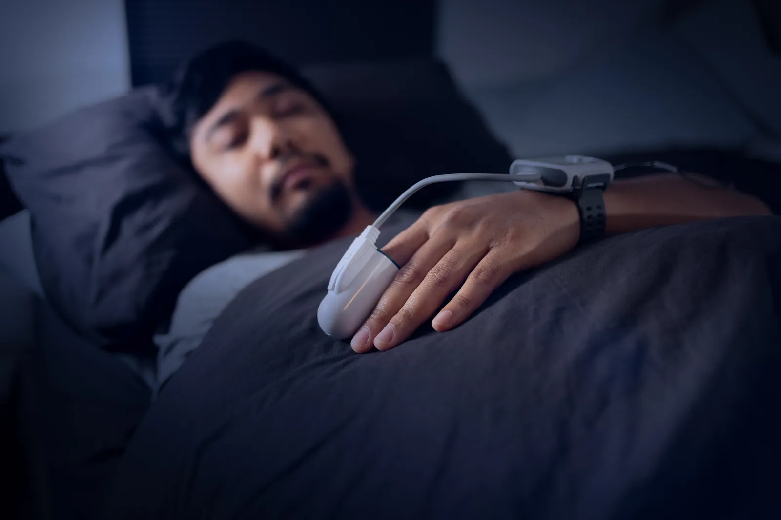 Man asleep in bed wearing a wrist-mounted sleep monitor with a finger sensor