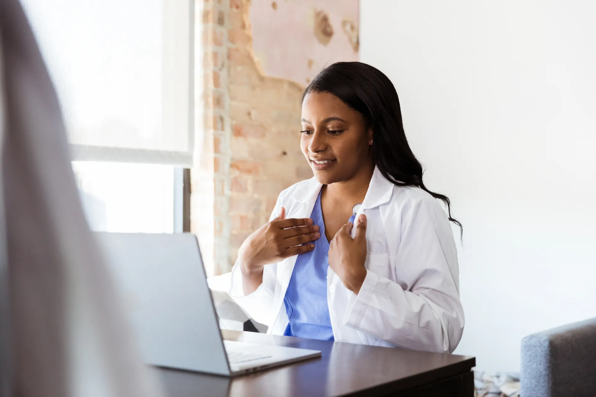 Smiling doctor in a white coat and scrubs talking to a patient on a laptop video call