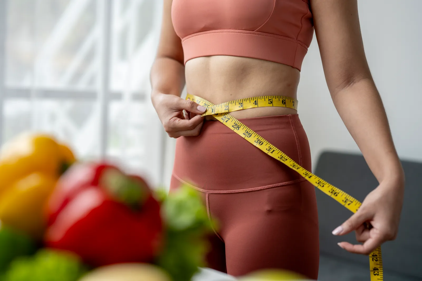 Woman in activewear measuring her waist with a tape measure behind blurred fresh vegetables