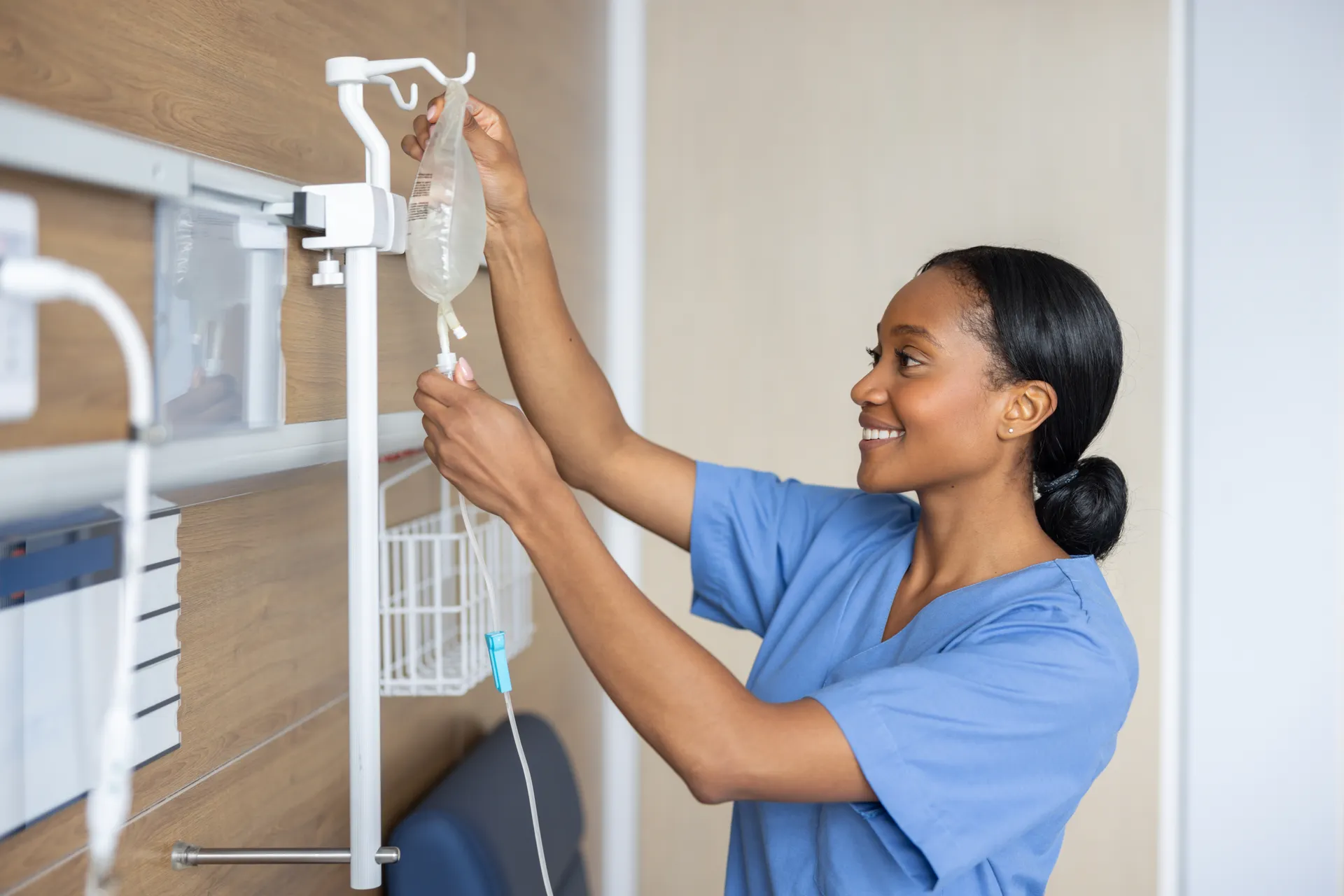 Smiling nurse in blue scrubs hangs an IV bag on a stand