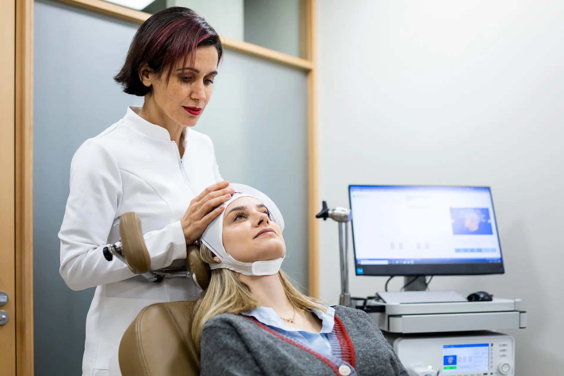 Clinician fitting a cap on a seated woman's head next to a TMS device and monitor