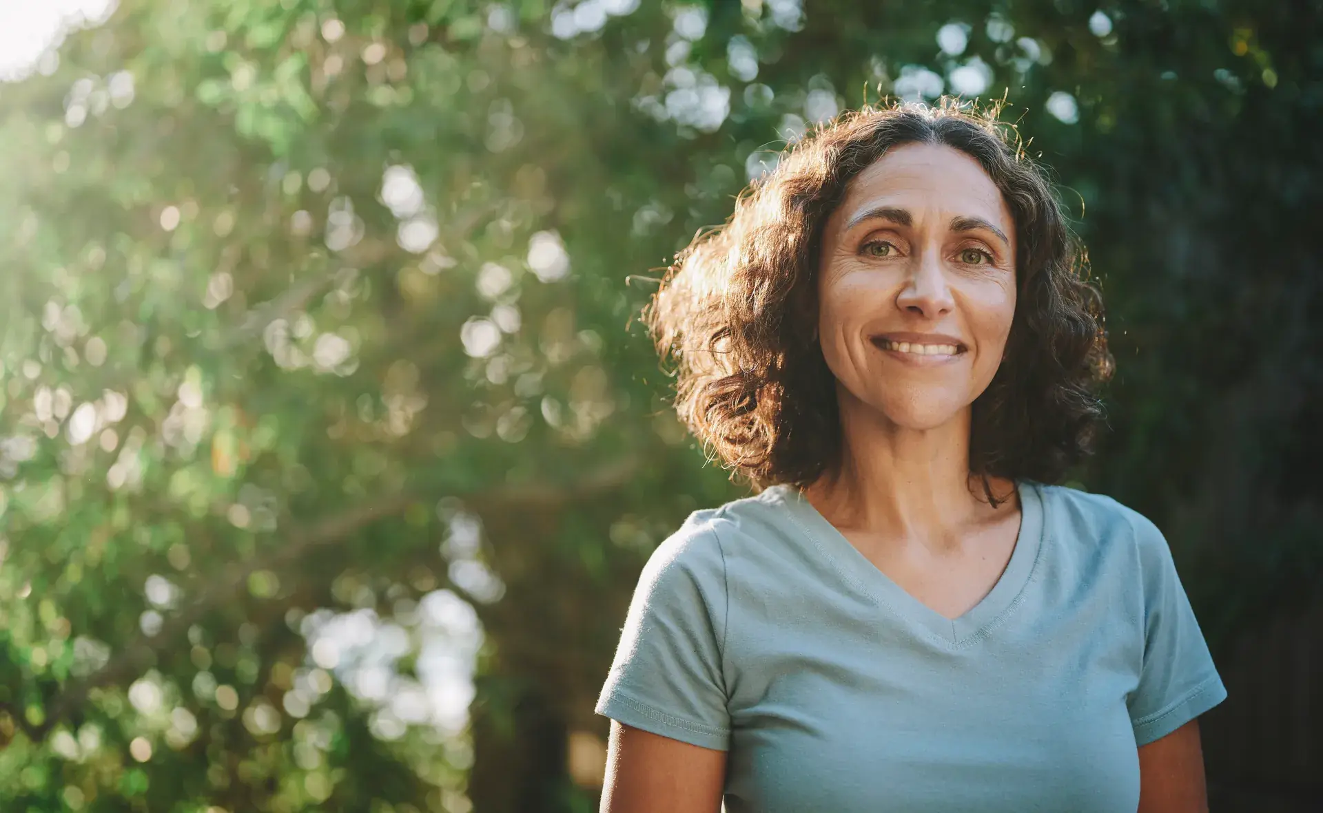 Smiling woman with curly hair standing outdoors in front of sunlit trees