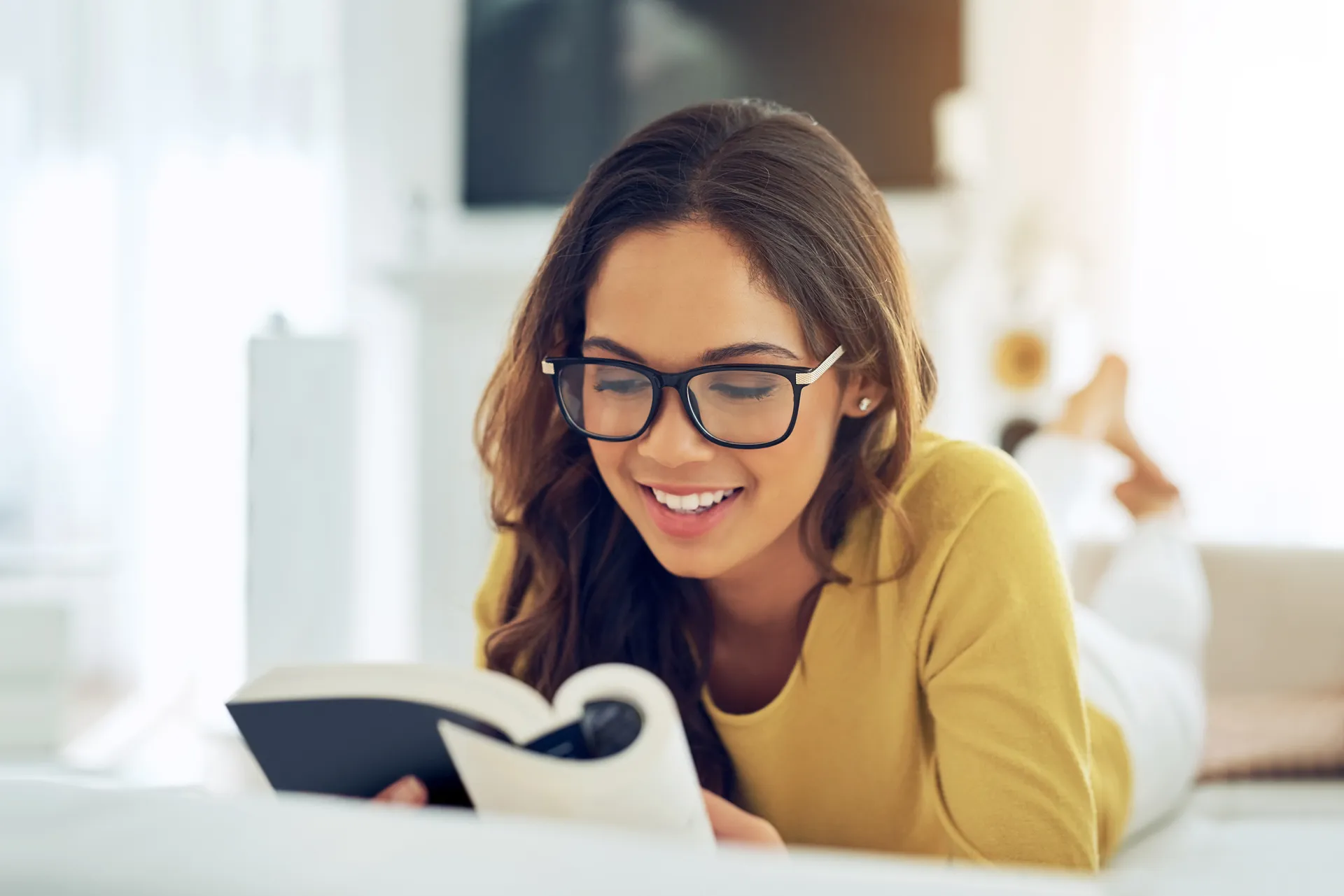Smiling young woman in glasses lying on a couch reading a book