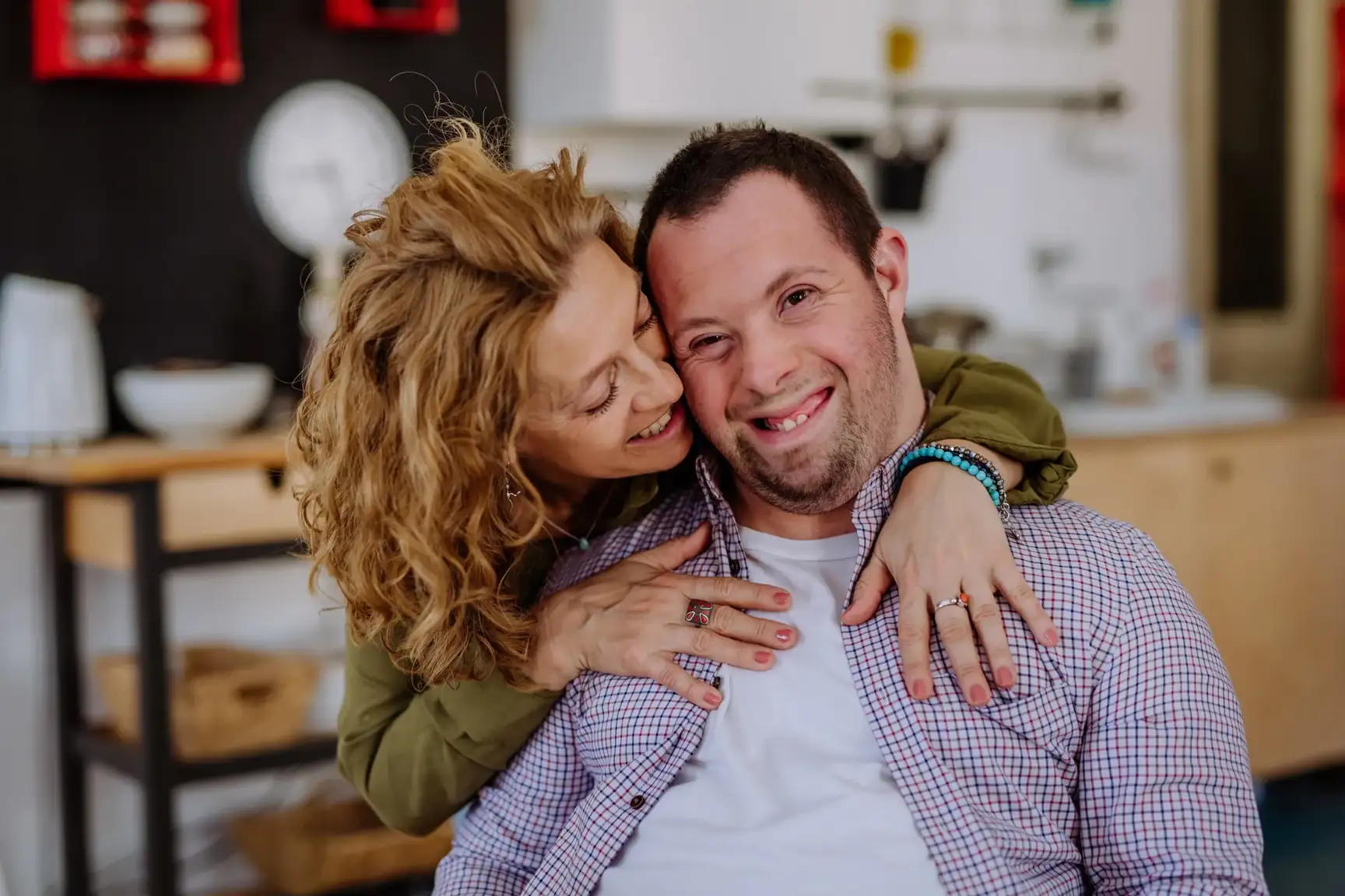 Smiling young man with Down syndrome being hugged from behind by a woman at home