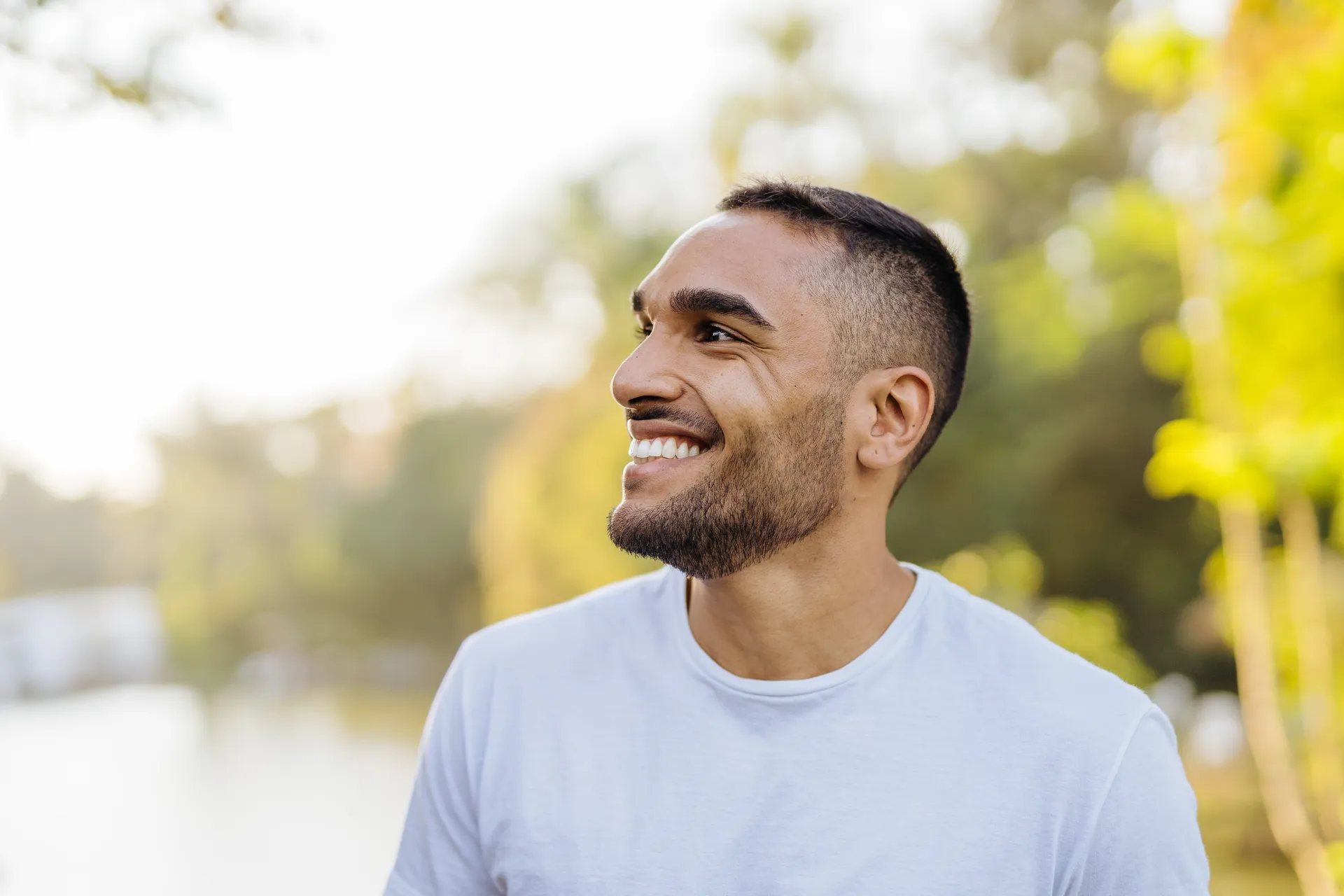 Smiling man in a white t-shirt outdoors with sunlit trees behind him