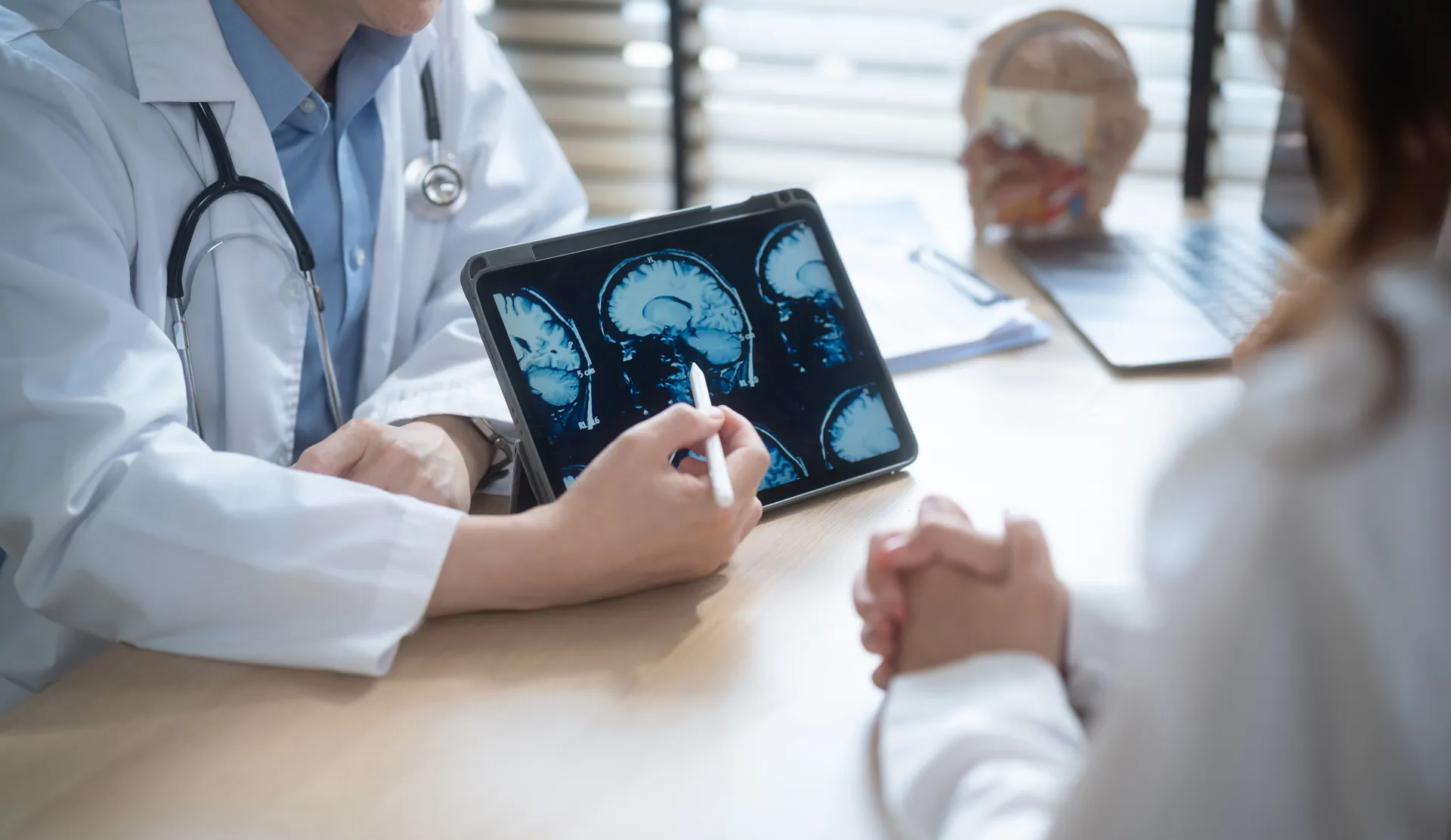 Doctor points a stylus at brain scan images on a tablet while talking with a patient across a desk