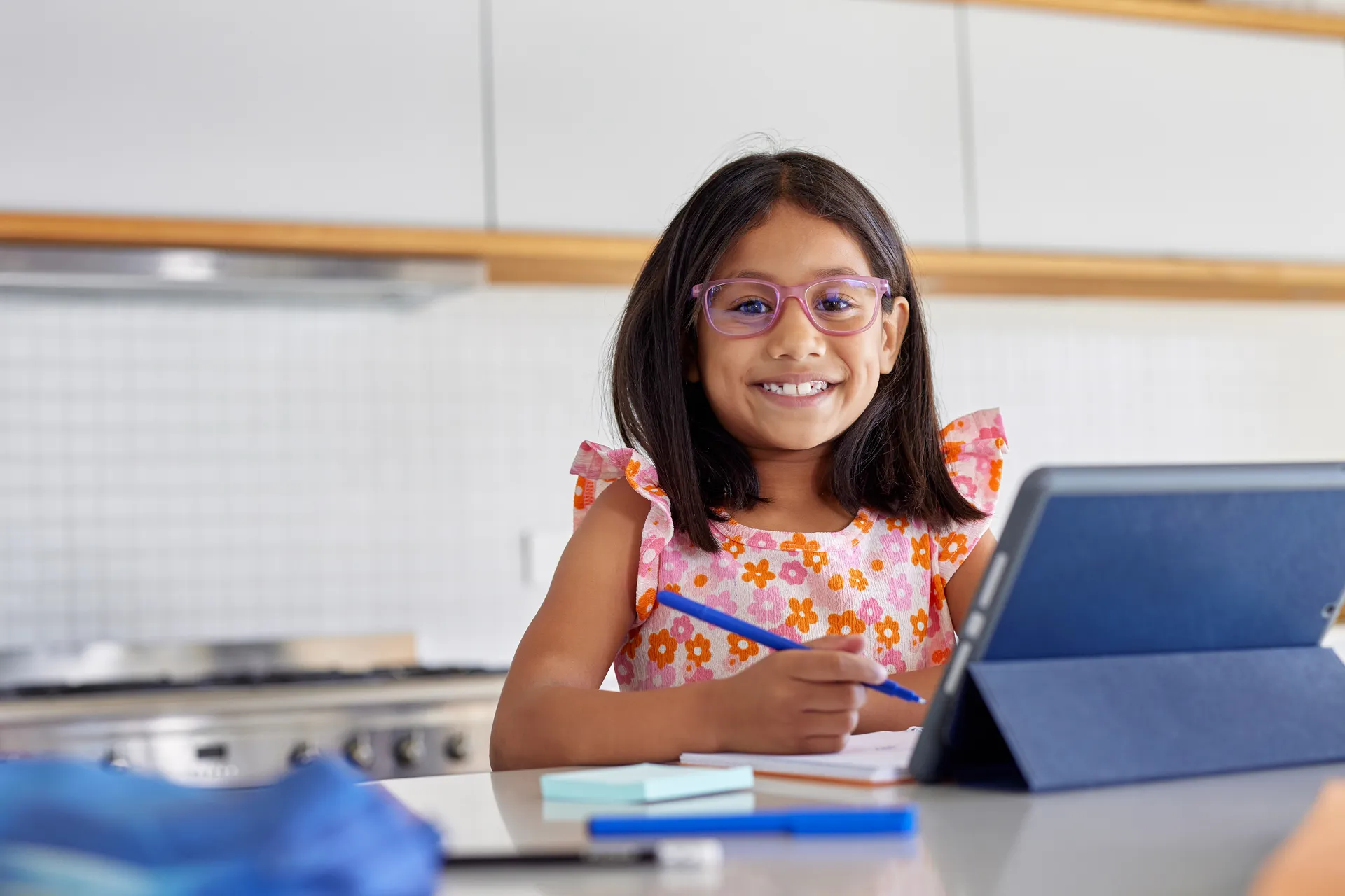 Smiling girl in glasses writing in a notebook beside a tablet at a kitchen counter