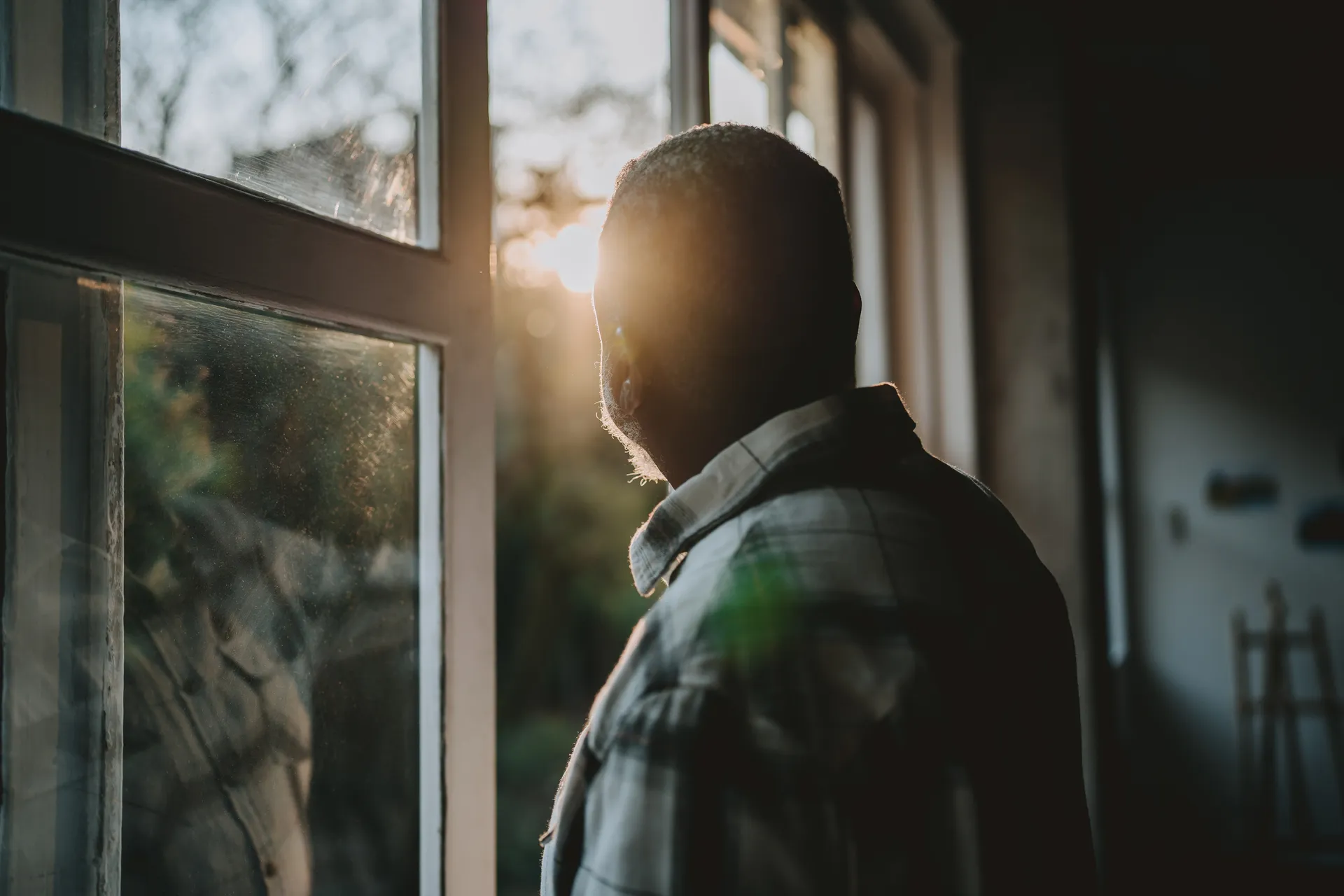Man in a plaid shirt stands in a dim room looking out a sunlit window