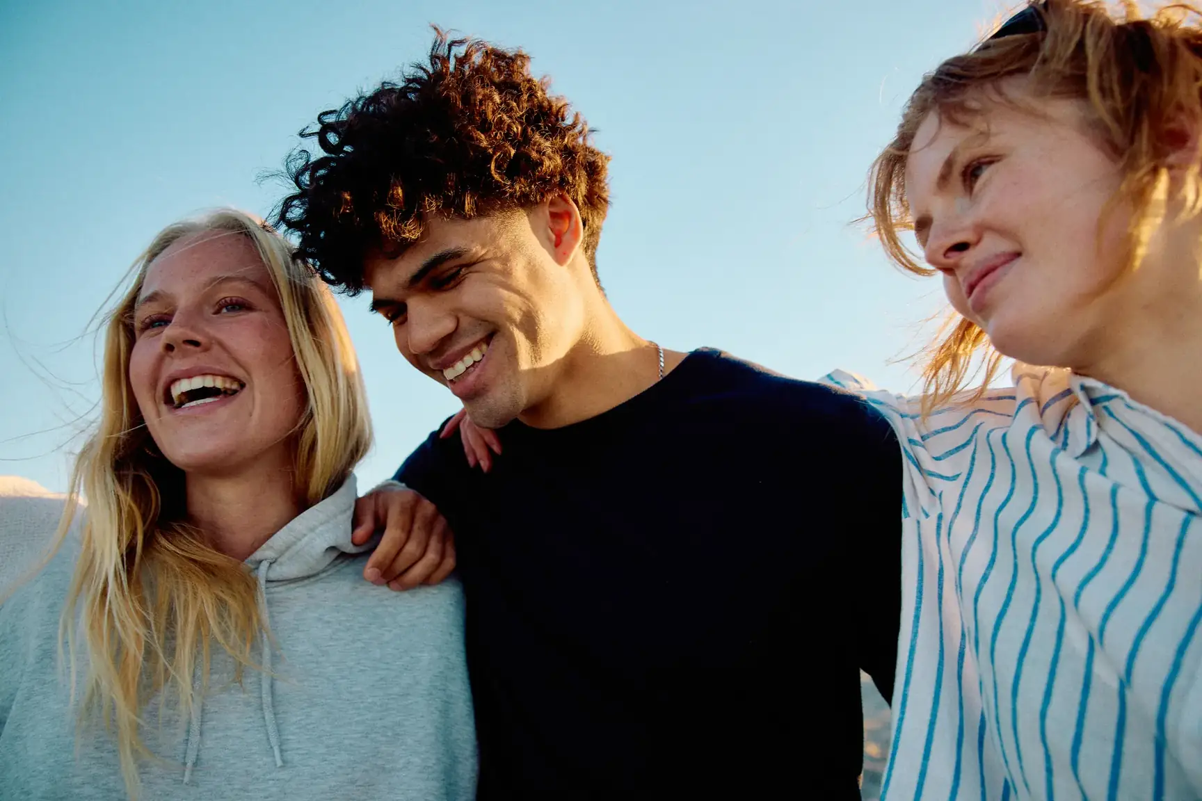 Three smiling friends with arms around each other outdoors under a blue sky