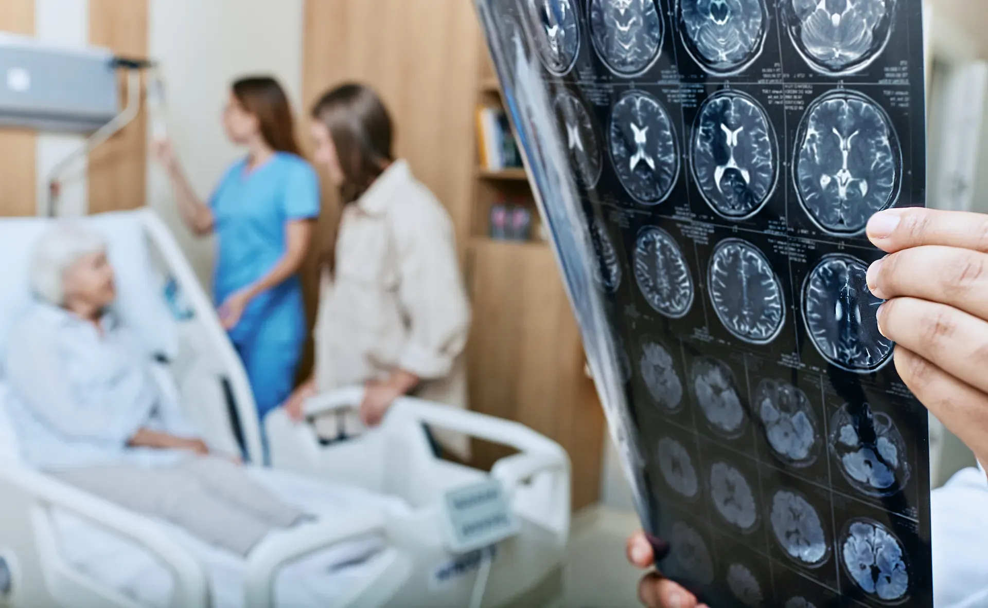 Clinician reviews brain scan films while an elderly patient in a hospital bed sits with a nurse and visitor
