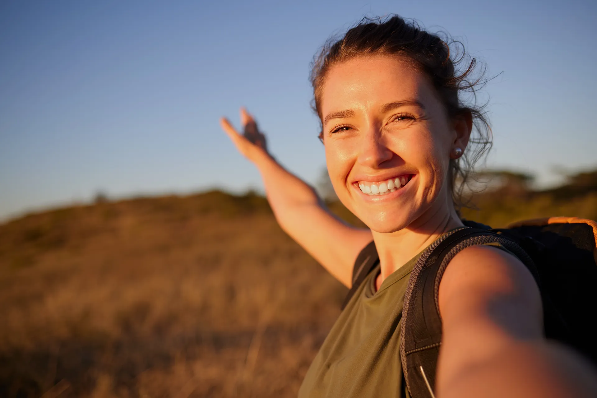 Smiling woman with a backpack taking a selfie on a hillside at sunset
