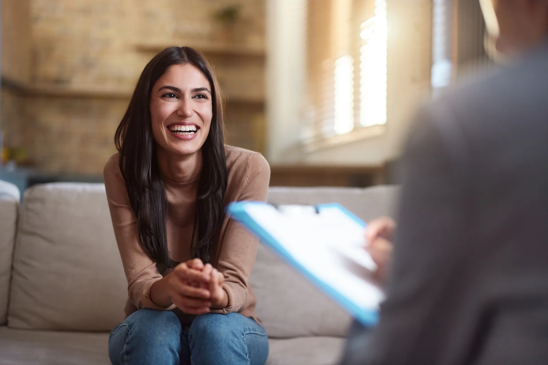 Smiling woman on a sofa talks with a therapist holding a clipboard