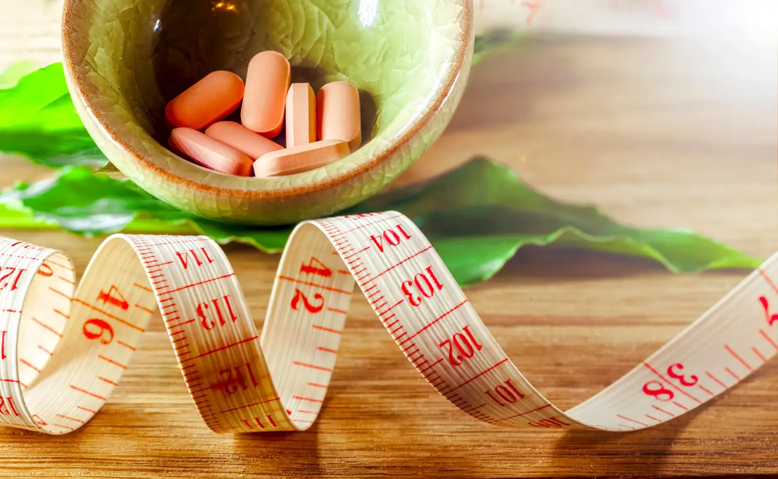 Bowl of pink tablets on green leaves with a measuring tape curled across a wooden table