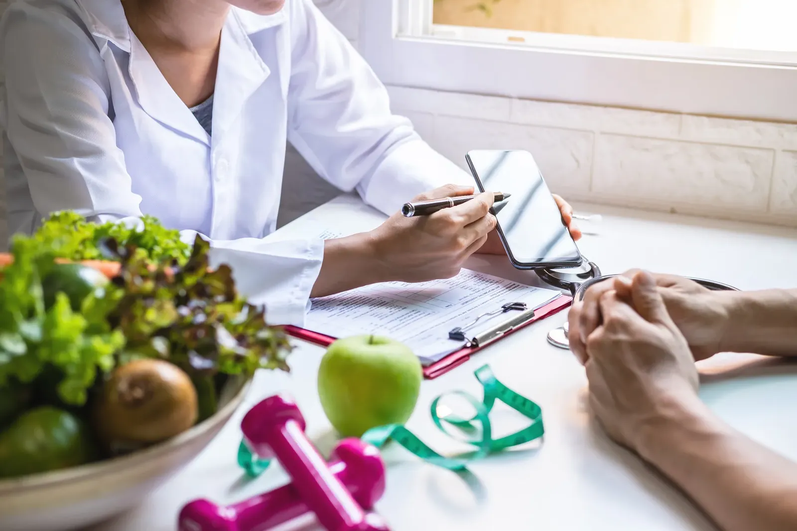 Clinician reviews notes with a patient beside a bowl of vegetables, an apple, dumbbells and a tape measure