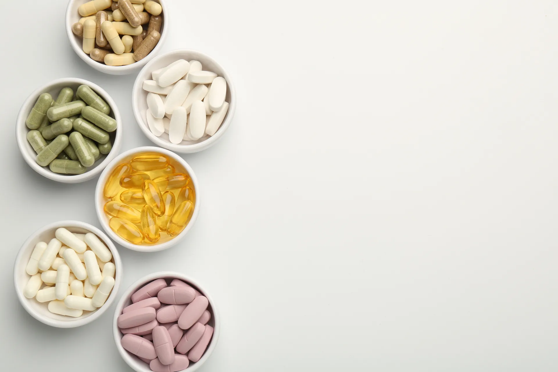 Small white bowls of assorted supplement capsules, softgels and tablets on a white surface