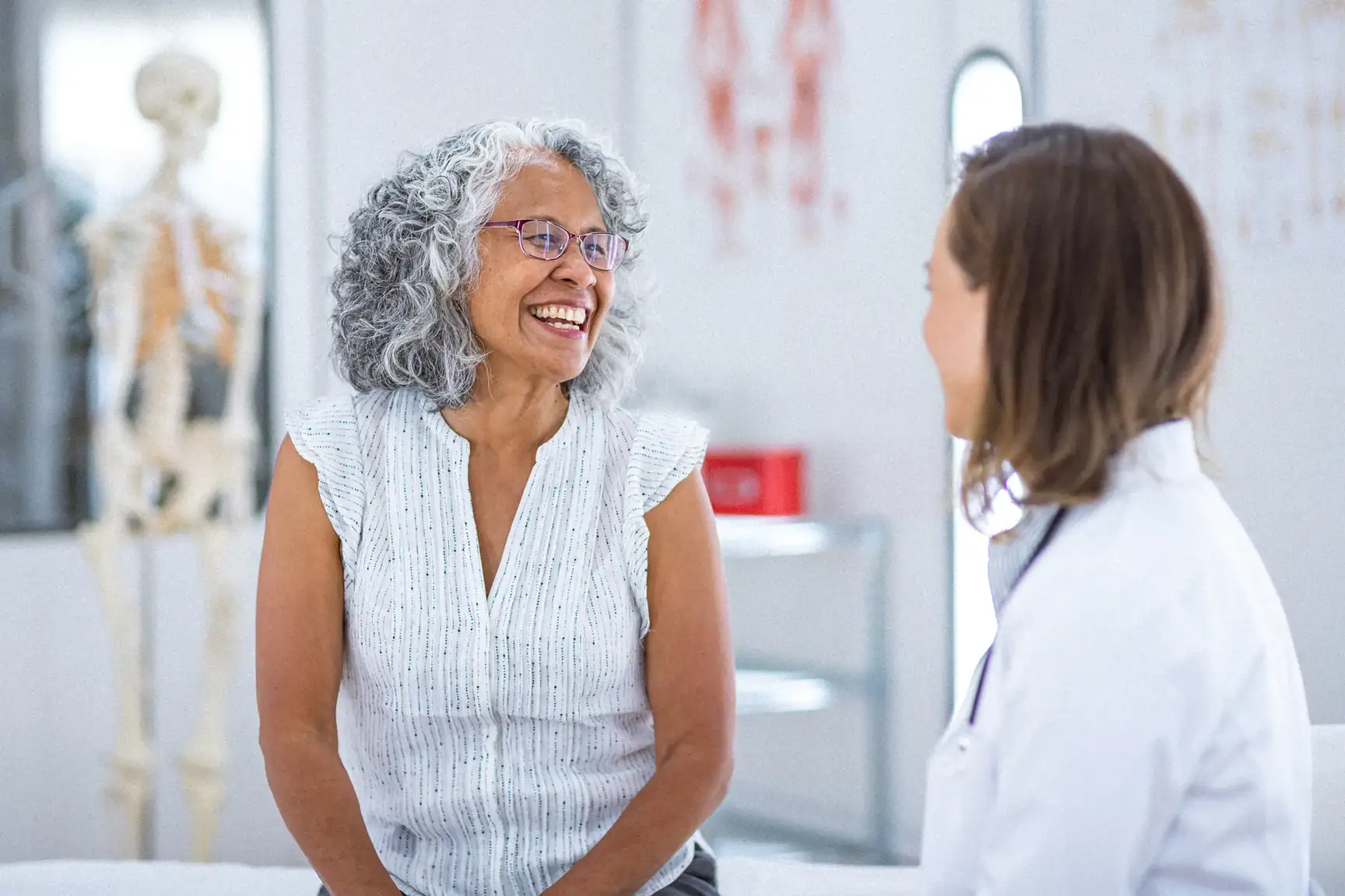 Smiling older woman with gray curly hair talks with a doctor in an exam room