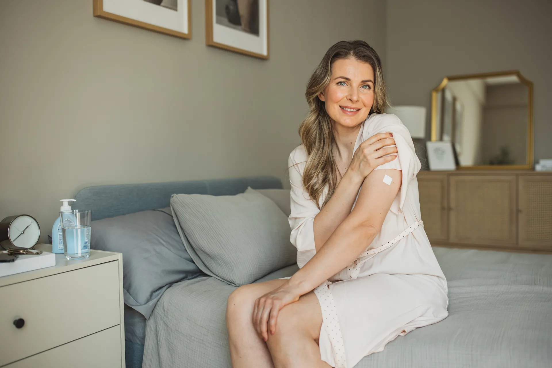 Smiling woman sitting on a bed holds her upper arm, which has a small bandage on it